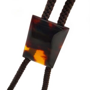 Op de afbeelding: Een bolo stropdas met een rechthoekige schuif met schildpadpatroon. De schuif is een mix van bruine, zwarte en amberkleuren. Het gevlochten koord is donkerbruin.