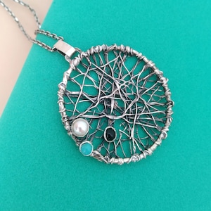 Puede incluir: Un collar con colgante plateado con un diseño circular en forma de telaraña. El colgante presenta una perla, una piedra turquesa y una piedra negra. El collar está sobre un fondo turquesa.
