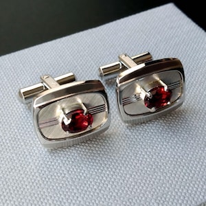 May include: A pair of rectangular silver-toned cufflinks featuring a brushed metal finish with horizontal etched lines. Each cufflink is centered with a single, oval-cut deep red gemstone held in place by a four-prong setting and a toggle back.