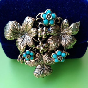 May include: Vintage gold-tone brooch featuring a leaf and grape design, accented with turquoise-colored beads. The brooch has a detailed, textured finish, and the turquoise beads add a pop of color to the piece.