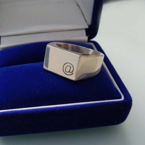 May include: A silver-colored ring with a rectangular face, featuring a black at symbol. The ring is displayed inside a blue velvet jewelry box with a white trim. The ring has a polished, reflective surface.