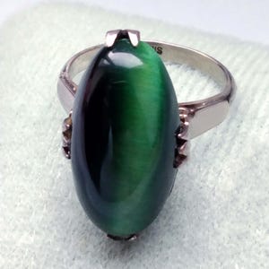 Puede incluir: Un anillo de plata con una gran piedra ojo de gato de color verde oscuro en forma ovalada, montada en un engaste de garras. La banda es lisa y metálica, con la palabra "SILVER" claramente grabada en la superficie interior, visible cerca del engaste.