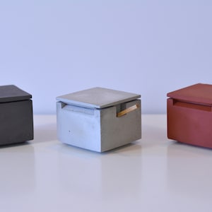 Peut inclure: Trois boîtes carrées en béton avec couvercles, en gris foncé, gris clair et rouge-brun. Chaque boîte a une petite ouverture rectangulaire et une fine poignée dorée. Les boîtes sont exposées sur une surface blanche.