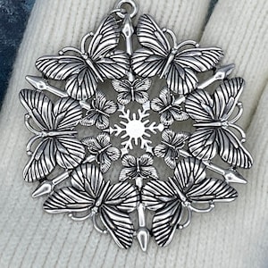 May include: A silver-toned metal pendant featuring a circular arrangement of six large butterflies surrounding a central snowflake-like star motif, with smaller butterflies interspersed between the larger ones and a small loop at the top.