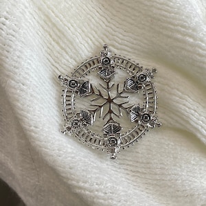 May include: A silver snowflake brooch with a detailed design. The snowflake is surrounded by a circular frame with intricate details.