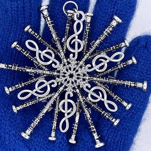 May include: A silver-coloured snowflake ornament featuring musical notes and clarinets. The ornament is designed with a central treble clef surrounded by multiple clarinets and additional treble clefs, creating a symmetrical, festive design. The ornament is held against a blue background.