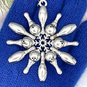 May include: A silver-toned metal ornament shaped like a snowflake, featuring eight bowling pins arranged in a radial pattern around a central star design. Each bowling pin has three small black dots near the top, and a small metal loop is attached to the top for hanging.