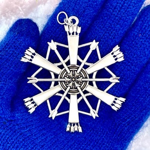 May include: A silver-toned metal pendant shaped like a snowflake, featuring a central circular hub with radiating arrow-like spokes. The design incorporates multiple arrow motifs pointing outward, creating a symmetrical, star-like geometric pattern.