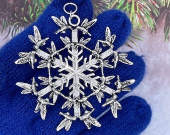 May include: A silver-toned metal ornament shaped like a snowflake, featuring intricate hummingbird figures arranged around a central star-like design. The ornament includes a small circular loop at the top for hanging and has a textured, metallic finish.