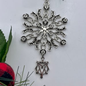 Peut inclure: Ornement de Noël en forme de flocon de neige argenté avec un motif de chardon et un charme suspendu représentant une couronne et la lettre "M".