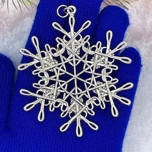 May include: A silver-toned metal snowflake ornament featuring a complex design composed of multiple small scissors arranged in a radial pattern. The ornament includes a circular loop at the top for hanging and has a polished, reflective metallic finish.