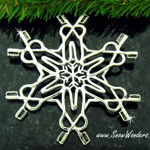 May include: A silver-toned metal snowflake ornament featuring a repeating pattern of six toothbrushes arranged in a radial design. The center of the ornament displays a small, intricate snowflake shape. The text www.SnowWonders.com is printed below.