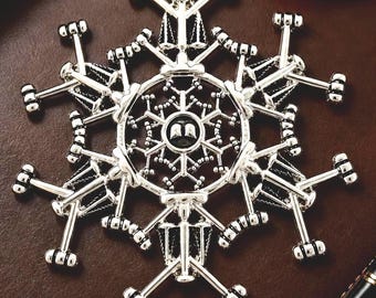 May include: A silver-toned metal ornament shaped like a snowflake, featuring intricate designs of small scales of justice and open books. The central circular medallion displays a pair of open books, with radiating arms ending in small hammer-like shapes.