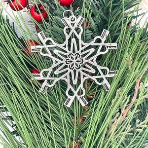 May include: A silver-toned metal snowflake ornament featuring a central star design surrounded by six radiating arms that resemble skeleton keys. The ornament includes a small circular loop at the top for hanging and has a polished metallic finish.