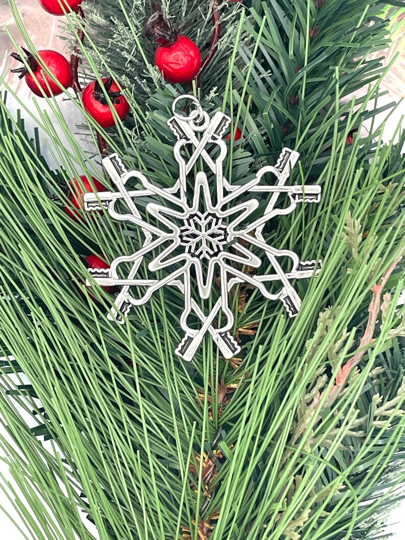 May include: A silver-toned metal snowflake ornament featuring a central star design surrounded by six radiating arms that resemble skeleton keys. The ornament includes a small circular loop at the top for hanging and has a polished metallic finish.