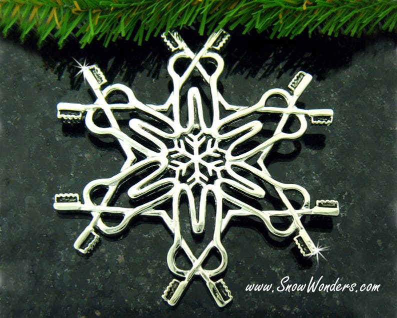 May include: A silver-toned metal snowflake ornament featuring a repeating pattern of six toothbrushes arranged in a radial design. The center of the ornament displays a small, intricate snowflake shape. The text www.SnowWonders.com is printed below.