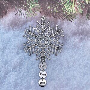 May include: A silver-toned snowflake ornament constructed from small bone shapes with a central paw print cutout. Three circular metal charms hang from the bottom, engraved with the names Rose and Zuma, and the year 2024 in a black sans-serif font.