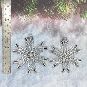 May include: Two silver-toned metal snowflake ornaments featuring a repeating pattern of crossed toothbrushes. A metal ruler is positioned on the left, showing the larger ornament measures approximately 3.5 inches in height and width.