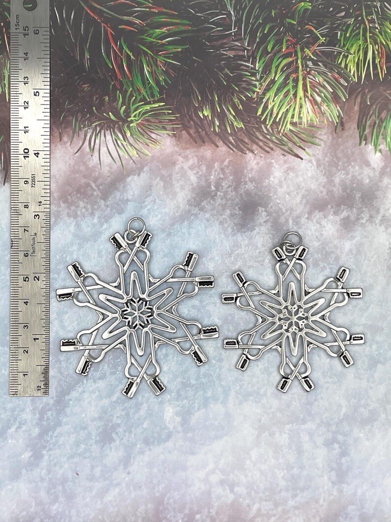 May include: Two silver-toned metal snowflake ornaments featuring a repeating pattern of crossed toothbrushes. A metal ruler is positioned on the left, showing the larger ornament measures approximately 3.5 inches in height and width.