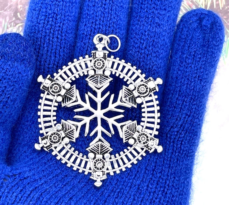 May include: A silver snowflake ornament with a train track design around the edge. Six small train engines are evenly spaced around the snowflake.