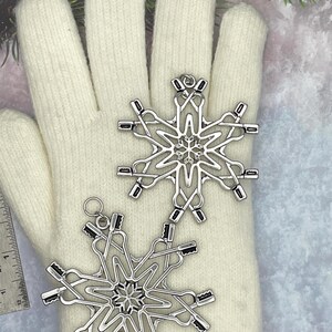 May include: Two silver-toned metal snowflake ornaments featuring a complex design of interlocking toothbrushes. Each ornament has a small circular loop at the top for hanging. A metal ruler is visible on the left, showing a scale in inches.