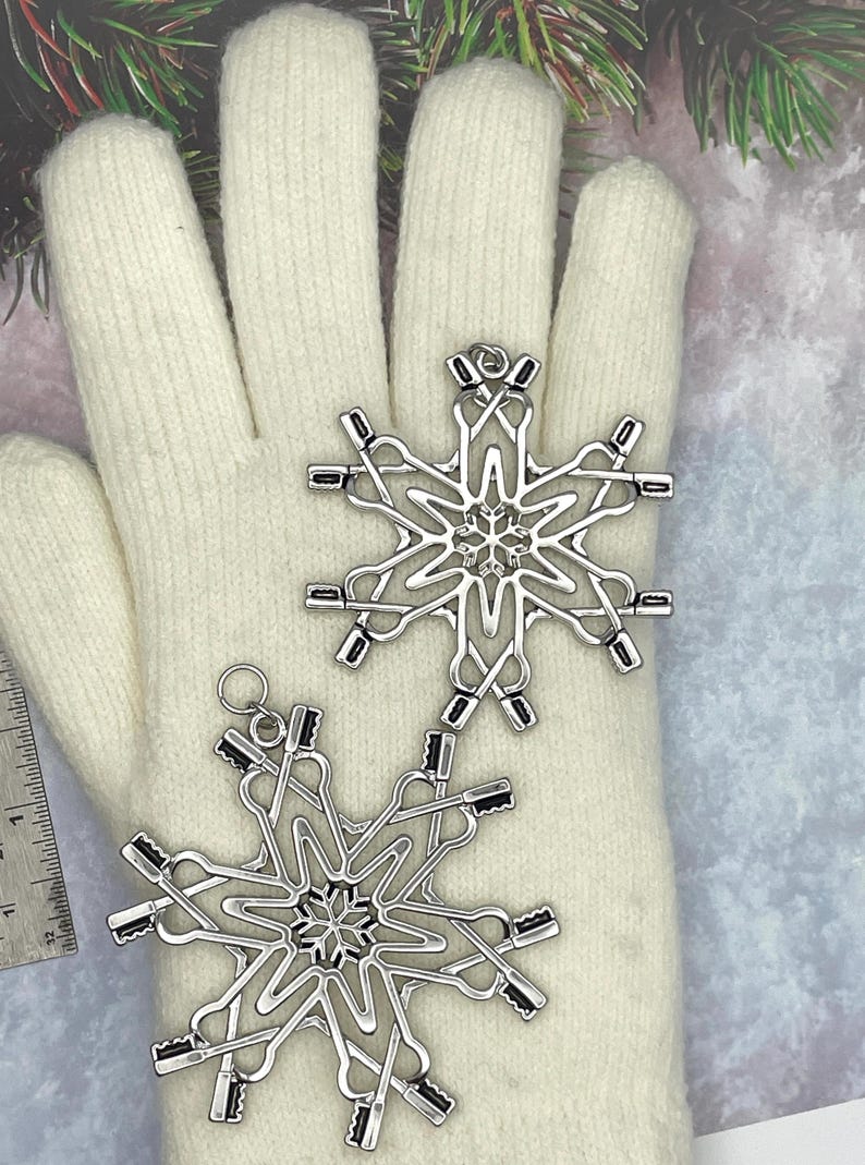 May include: Two silver-toned metal snowflake ornaments featuring a complex design of interlocking toothbrushes. Each ornament has a small circular loop at the top for hanging. A metal ruler is visible on the left, showing a scale in inches.