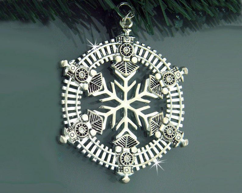 May include: Silver snowflake Christmas ornament with a train track design around the edges. Six small train engines are positioned around the snowflake.