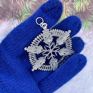 May include: A silver snowflake ornament with a detailed design. The ornament is held in a blue knitted glove.