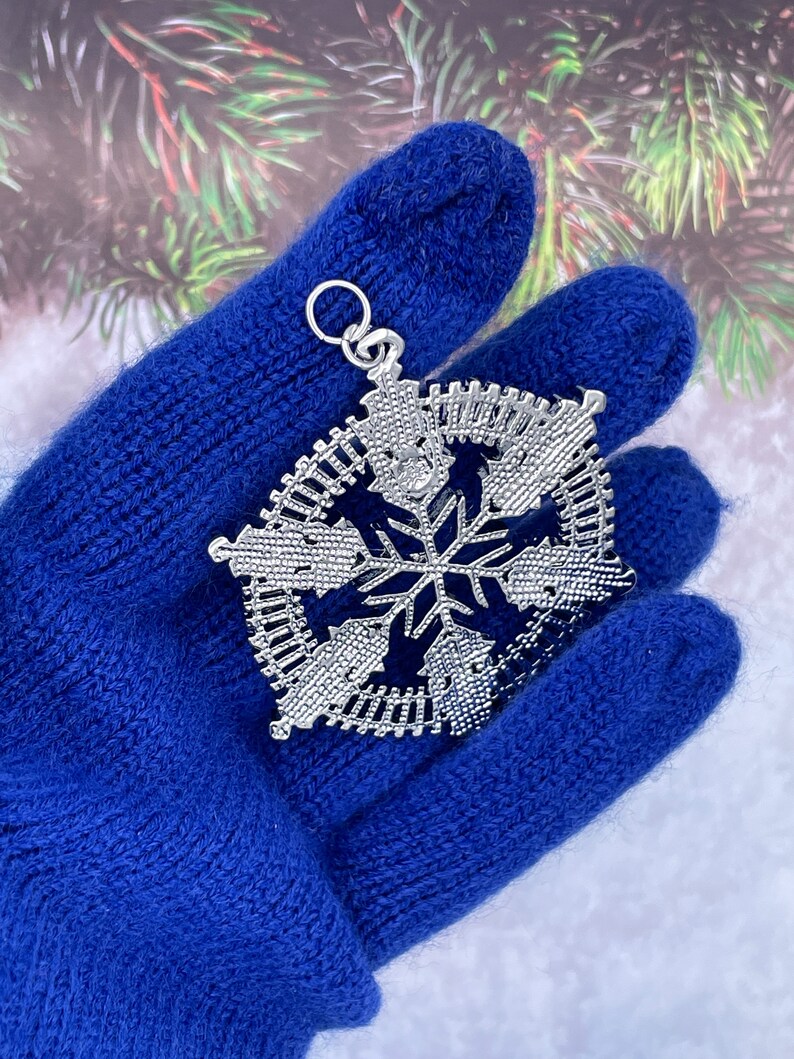 May include: A silver snowflake ornament with a detailed design. The ornament is held in a blue knitted glove.