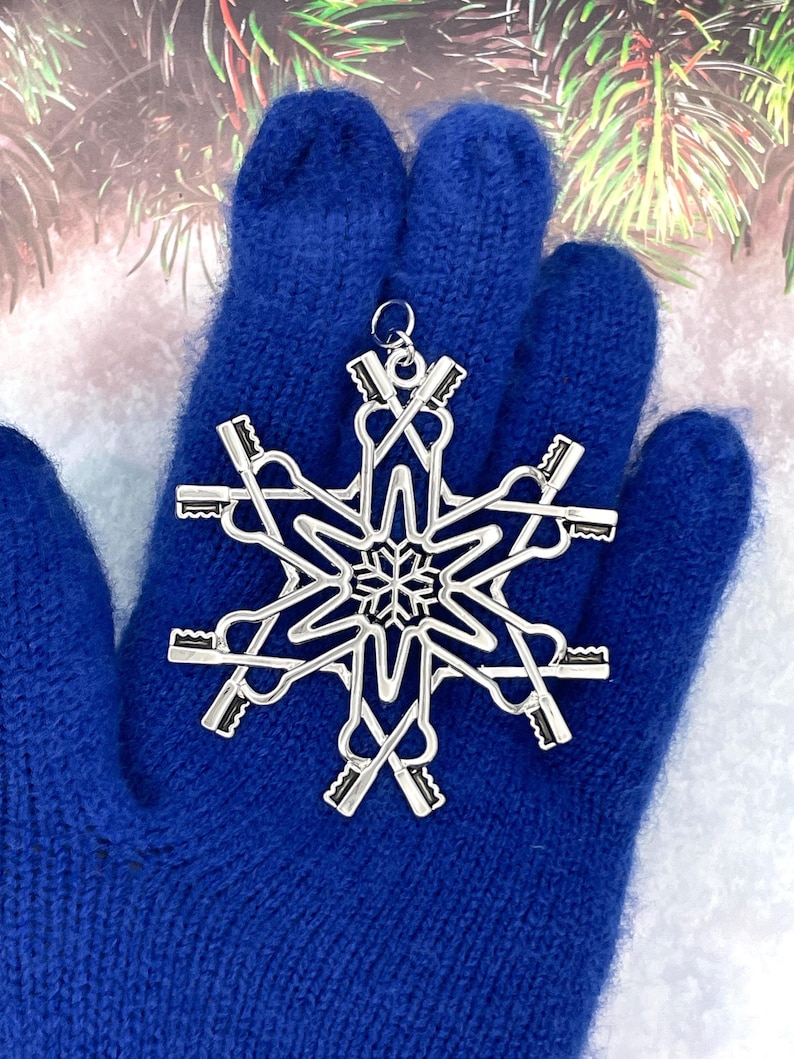 May include: A silver-toned metal ornament shaped like a snowflake, constructed from multiple small key silhouettes arranged in a radial pattern. The center features a detailed snowflake cutout, and a small circular loop is attached at the top.