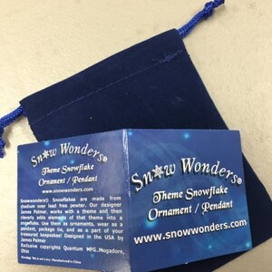May include: A dark blue velvet drawstring pouch accompanied by a folded informational card. The card features the text Snow Wonders Theme Snowflake Ornament / Pendant and includes details about the product design and manufacturing origins.