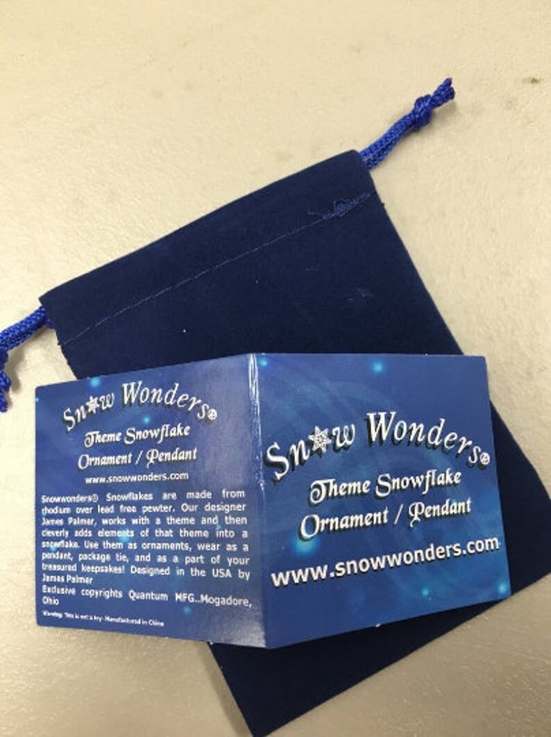 May include: A dark blue velvet drawstring pouch accompanied by a folded informational card. The card features the text Snow Wonders Theme Snowflake Ornament / Pendant and includes details about the product design and manufacturing origins.