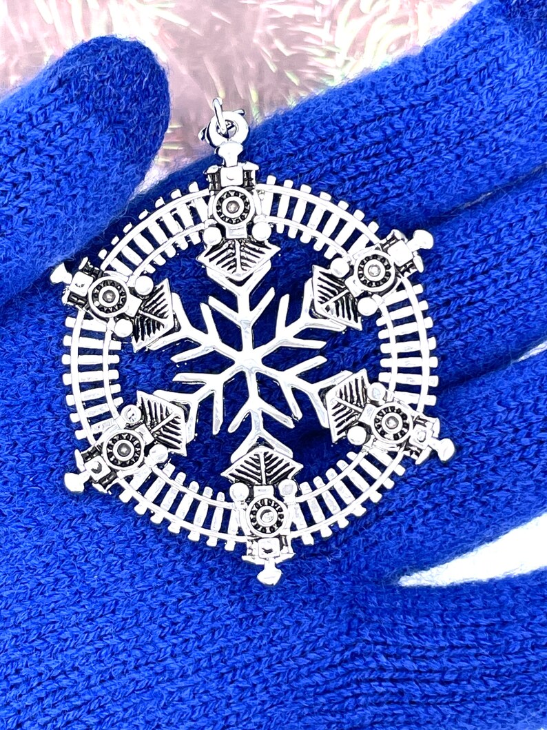 May include: Silver snowflake Christmas ornament with a train track design and six small train engines around the snowflake.