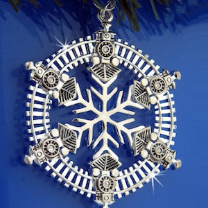 May include: A silver snowflake Christmas ornament with a train track design and six miniature train engines around the perimeter.