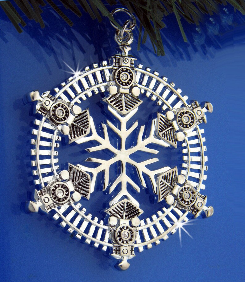 May include: A silver snowflake Christmas ornament with a train track design and six miniature train engines around the perimeter.