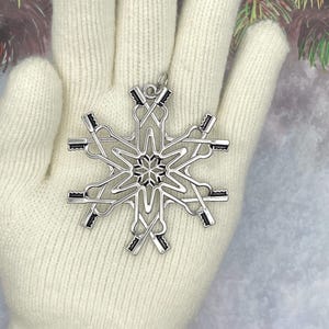 May include: A silver-toned metal snowflake ornament featuring a repeating pattern of crossed rowing oars. The design includes a small circular loop at the top for hanging, with a central star-like detail and six symmetrical arms extending outward.
