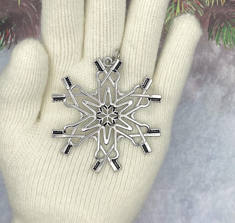 May include: A silver-toned metal snowflake ornament featuring a repeating pattern of crossed rowing oars. The design includes a small circular loop at the top for hanging, with a central star-like detail and six symmetrical arms extending outward.