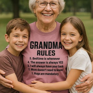 May include: A smiling woman wearing glasses hugs two children. The woman's mauve shirt reads "GRANDMA RULES" with a list of rules below. The children are smiling and wearing casual clothing. The background is a blurred green outdoor scene.