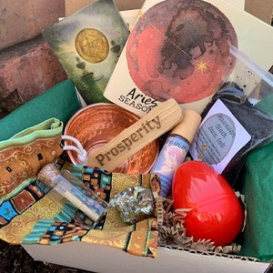 Puede incluir: Una caja blanca llena de varios artículos para un ritual o hechizo. Los artículos incluyen un pequeño cuenco de cobre, un palo de madera con la palabra "Prosperity" grabada en él, una pequeña botella con un tapón de corcho, una pequeña bolsa de sal de lava negra, un huevo rojo y una tarjeta con las palabras "Aries Season" en ella.