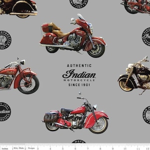 Op de afbeelding: Een grijze stof met een herhalend patroon van vintage Indian-motorfietsen in rood en zwart, afgewisseld met cirkelvormige logo's. De tekst luidt AUTHENTIC Indian MOTORCYCLE SINCE 1901. Een witte liniaal onderaan toont afmetingen in centimeters.