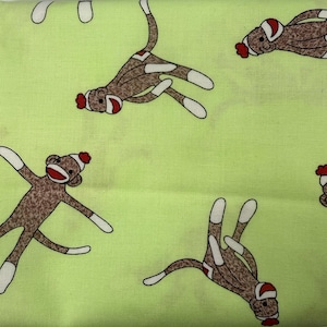 May include: A light green fabric featuring a repeating pattern of brown sock monkeys. Each monkey has white accents on the hands, feet, and face, along with a red mouth and a red pom-pom on the top of its head. The monkeys are oriented in various directions.