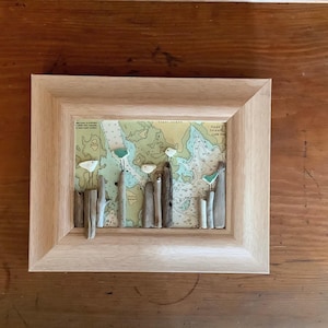May include: A framed artwork featuring a map background with driftwood and small white and teal accents. The frame is made of light-colored wood. The artwork has a nautical theme.