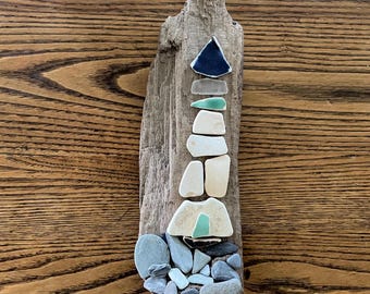 May include: A driftwood lighthouse sculpture. The lighthouse is constructed from various pieces of sea glass and small stones in shades of blue, white, and gray. The base is made of small, rounded stones, and the tower is built from stacked pieces of sea glass.