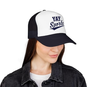 May include: A navy blue and white trucker hat with the text "YAY Sports" in navy blue script. The hat has a mesh back and a curved brim. The hat is a baseball cap style.