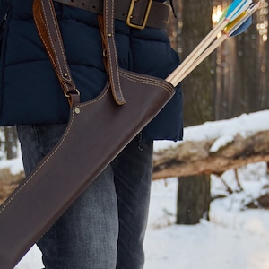 May include: A brown leather quiver with a strap and buckle, worn over a blue jacket and jeans. The quiver is filled with arrows, and the person is standing in a snowy forest.