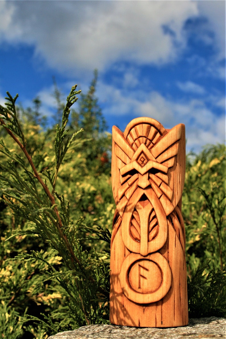 May include: A wooden carving of a Norse god with intricate details. The carving is painted in shades of brown and features a rune symbol. The carving is set against a backdrop of green foliage and a blue sky.