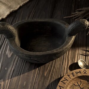 May include: A dark wooden bowl with two raven heads as handles. The bowl is sitting on a wooden table with a wooden plate with runes on it.