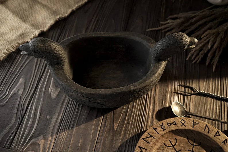 May include: A dark wooden bowl with two raven heads as handles. The bowl is sitting on a wooden table with a wooden plate with runes on it.