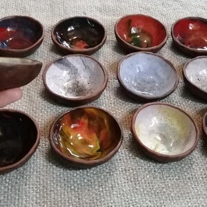 May include: A collection of 12 small ceramic bowls with various colorful glazes. The bowls are arranged in a semi-circle on a textured surface. Some bowls have a speckled glaze, while others have a more solid color.