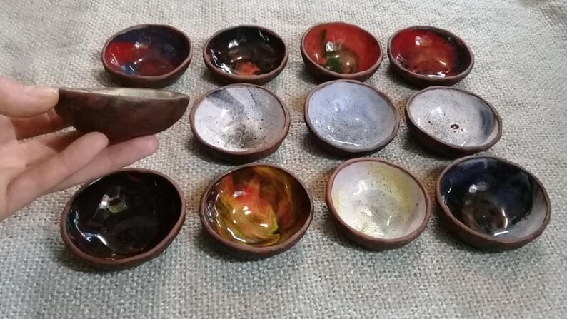 May include: A collection of 12 small ceramic bowls with various colorful glazes. The bowls are arranged in a semi-circle on a textured surface. Some bowls have a speckled glaze, while others have a more solid color.